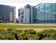 Moderne Bürogebäude mit großen Glasfassaden und weißen Fassadenelementen, umgeben von einer Grünfläche mit niedrigem Gebüsch im Vordergrund. Im Hintergrund sind ein weiteres mehrstöckiges Bürogebäude mit einem „Oracle“-Schriftzug und einige Passanten zu sehen.