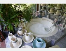 Ein Badbereich mit einer eingelassenen, runden Badewanne, die mit Schaumwasser gefüllt ist. Rundherum stehen dekorative Porzellankannen, Schalen und Vasen, sowie Topfpflanzen neben einem Fenster mit Blick ins Grüne.