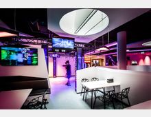 Ein moderner Unterhaltungsbereich mit futuristischer Einrichtung, darunter Tische mit geometrischen Stühlen und beleuchtete Wandelemente. Im Hintergrund ist eine Virtual-Reality-Station mit Bildschirmen und schwebenden Kabeln zu sehen, beleuchtet von bunten, lila- und pinkfarbenen Neonlichtern.