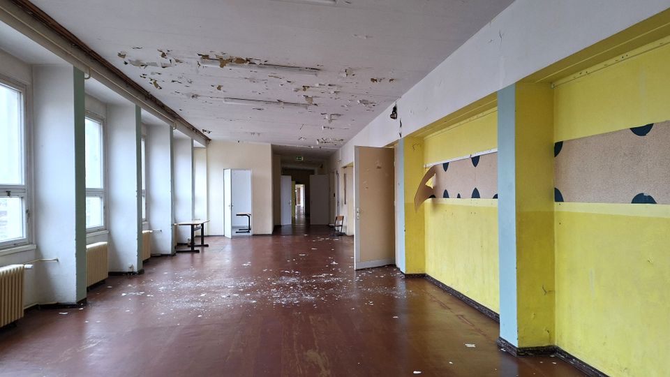 Langer, leerer Flur in einem älteren Gebäude mit großen Fenstern und Heizkörpern auf der linken Seite sowie mehreren Türen entlang des Gangs. Die Decke zeigt abblätternde Farbe und der Boden ist mit Farbresten und Schmutzspuren bedeckt; rechts sind gelb gestrichene Wände mit verblasster Wanddekoration zu sehen.