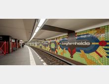 U-Bahn-Station mit dem Namen „Jungfernheide“, gekennzeichnet durch bunte, geometrische Mosaikmuster an der Wand. Die Station hat eine leicht gekrümmte Bahnsteigkante mit Schienen in der Mitte und modernem weißem Deckenlicht.