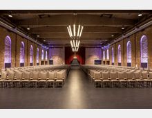 Großer Veranstaltungsraum mit Reihen von beigen Stühlen, einer Bühne mit geschlossenem roten Vorhang im Hintergrund und Backsteinwänden mit violett beleuchteten Rundbogenfenstern. Die Decke besteht aus freiliegenden Balken und moderne Hängelampen dominieren die Mitte des Raumes.