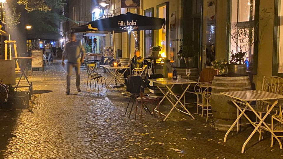 Nächtliche Szene einer schmalen Kopfsteinpflasterstraße mit Außengastronomie: Klapptische und Stühle stehen entlang einer Gebäudefassade unter großen Sonnenschirmen, beleuchtet von Laternen und warmem Innenlicht aus Fenstern. Pflanzkübel und Dekoelemente säumen den Rand, im Hintergrund setzt sich die Straße mit weiteren Leuchten und Sitzbereichen fort.