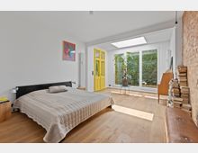 Helles Schlafzimmer mit Holzboden, einem niedrigen Bett mit beigefarbener Tagesdecke, einer gelben Tür und großen Fenstern, die zu einem begrünten Außenbereich führen. An der rechten Wand aus Ziegelsteinen steht ein Bücherstapel; ein Oberlicht lässt zusätzliches Tageslicht herein.