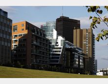 Moderne städtische Skyline mit mehreren Gebäuden in verschiedenen Architekturstilen, darunter zeitgenössische Glasfassaden und Backsteinfassaden. Im Vordergrund befindet sich eine offene Grünfläche, umgeben von Bäumen.