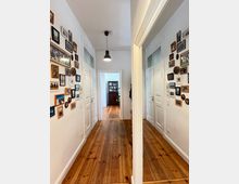 Ein heller Flur mit Holzdielenboden und weißen Wänden, geschmückt mit zahlreichen gerahmten Fotos an den Seitenwänden. Am Ende des Flurs ist ein Raum mit einem Holzschrank sichtbar, während eine Hängeleuchte die Decke ziert.