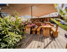 Biergartenähnliche Außenterrasse auf einem Holzdeck mit rustikalen Holztischen und -bänken, teils mit rot-weiß karierten Tischdecken, unter großen Stoff-Sonnensegeln. Im Hintergrund stehen eine Holzbar und Pflanzenkübel, dahinter eine bemalte Wand und moderne Bürogebäude.
