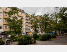 Wohnhausfassaden an einem Platz mit Bäumen und Sträuchern, darunter links ein gelbes Gebäude mit Balkonen und rechts ein Wohnhaus mit bunten, künstlerisch bemalten Balkonen. Im Vordergrund befinden sich Sitzbänke, Tischgruppen und Gehwege.