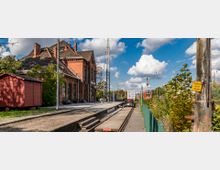 Ein kleiner Bahnhof mit einem historischen Backsteingebäude, daneben ein rotes Holzhäuschen. Im Vordergrund verläuft ein Gleis, im Hintergrund sind Bahnanlagen und Vegetation zu sehen, unter einem teils bewölkten Himmel.