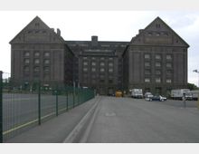 Backsteinbau eines alten Lagerhauses mit der Aufschrift „Zollspeicher“ auf der linken und „Westhafen“ auf der rechten Giebelseite. Im Vordergrund befinden sich ein umzäunter Vorplatz, parkende Lkws und Autos, sowie ein Asphaltweg, der zu dem Gebäude führt.