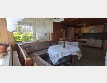 Gemütliche Küche mit Holzwänden und Fliesenboden. Zu sehen sind eine Eckbank mit Tisch, dekoriert mit einer Tischdecke und Blumenvase, ein großes Fenster mit Spitzengardinen sowie eine Einbauküche mit weißen Schränken und Küchengeräten.