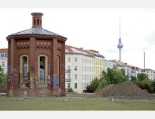 Backsteinbau mit oktogonalem Grundriss und kleinen Fenstern, teilweise mit Graffiti bedeckt, im Vordergrund einer Grünfläche mit aufgeschüttetem Erdwall und Bauzaun; im Hintergrund Wohnhäuser und der Berliner Fernsehturm.