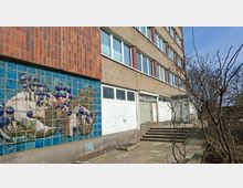 Außenansicht eines mehrstöckigen Wohn- oder Bürogebäudes mit rau verputzter Fassade und langen Fensterreihen. Im Vordergrund ein Eingangsbereich mit Treppenstufen und Gehweg, daneben eine große blaue Fliesenwand mit einem floralen Mosaik sowie kahle Sträucher.