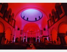 Innenraum einer Kirche mit beleuchteter Kuppeldecke, die in kräftigem Rot und Blau erstrahlt. Rundbögen und Säulen prägen die Architektur, im Hintergrund ist ein großes Kreuz über dem Altar sichtbar.