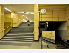 Innenansicht einer U-Bahn-Station mit gelb gefliesten Wänden, einer breiten Treppe, die nach oben führt, und einem Bahnsteig auf der rechten Seite. Eine analoge Uhr ist an der Wand angebracht, und ein Spiegel reflektiert das Gleis und die Umgebung.