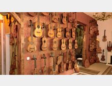 Ein Raum mit einer Sammlung von Akustikgitarren, die dicht an einer roten, verzierten Wand aufgehängt sind. Im Hintergrund sind ein Kamin, ein Kronleuchter sowie weitere Musikinstrumente erkennbar.