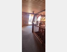 Ein Dachgeschossraum mit holzvertäfelten Wänden und Decke sowie Fenstern, die viel Tageslicht hereinlassen. Im Vordergrund befindet sich ein Geländer, und im Hintergrund ist eine Glastür, die zu einem Balkon führt.