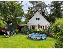 Einfamilienhaus mit steilem Satteldach und weißen Fassaden, umgeben von dichtem Baumbestand und Gartensträuchern. Im Vordergrund liegt eine gepflegte Rasenfläche mit rundem Aufstellpool, Liegestühlen und einem Trampolin am Rand.