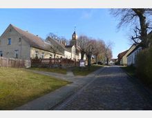Kopfsteinpflasterstraße in einem Dorf mit historischen Gebäuden, darunter ein Haus mit Solarzellen und eine kleine Kirche mit Turmuhr im Hintergrund; entlang der Straße stehen Laternen und Bäume ohne Laub.