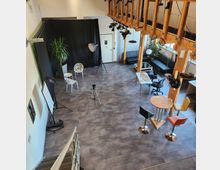 Großer, offener Raum mit hellgrauem Boden, eingerichtet als Fotostudio und Arbeitsbereich. Links befindet sich eine schwarze Fotowand mit Stühlen, Pflanzen und Stativen; rechts sind Tische, Stühle und Sofas angeordnet. Holzbalken und Industrieleuchten prägen die Architektur.