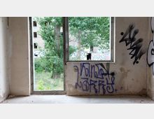 Blick aus einem verlassenen, teils verfallenen Raum mit Graffiti auf den Wänden und einem großen offenen Fensterrahmen. Draußen sind Bäume und ein weiteres Gebäude mit Graffiti sichtbar.
