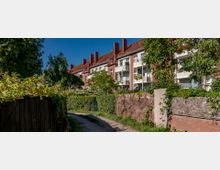 Mehrfamilienhäuser im Stil der Backsteinarchitektur mit roten Ziegeldächern und kleinen Balkonen, umgeben von Bäumen, Büschen und einer gepflasterten Gehwegspur, die durch eine grüne Wohnanlage führt.