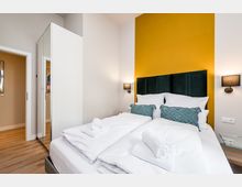 Modernes Schlafzimmer mit Doppelbett, weißer Bettwäsche und gerollten Handtüchern vor einer senfgelben Akzentwand mit gepolstertem dunklem Kopfteil. Seitlich stehen ein hoher weißer Kleiderschrank mit Spiegeltür, zwei Wandlampen und ein kleiner Glastisch auf Holzboden.