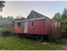 Ein roter, hölzerner Bauwagen mit einem gewölbten Dach steht auf einer Wiese neben einem Holzzaun und einem Gebäude mit Ziegeldach. Im Hintergrund sind Bäume und Buschwerk sichtbar, die eine ländliche Umgebung andeuten.