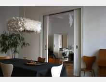 Heller Essbereich mit großem Tisch und Stühlen, einer federartigen Hängelampe und hohen weißen Wänden. Durch eine breite Schiebetüröffnung ist ein angrenzendes Zimmer mit Bücherstapel auf einem Regal, Teppichboden und geöffneten Türen zu sehen.