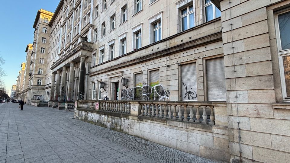 Lange, mehrstöckige Sandsteinfassade eines historischen Gebäudekomplexes entlang eines breiten Gehwegs, mit hohen Säulen am Eingangsbereich. Im Erdgeschoss sind mehrere Fenster mit Rollläden sowie Graffiti zu sehen, davor eine niedrige Balustrade aus Stein.