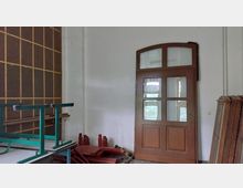 Ein Raum mit weißen Wänden und einer großen, braunen Holztür mit Fenstereinsätzen. Im Vordergrund Stapel von Holz- und Metallobjekten sowie eine Wand mit einem Gittermuster auf der linken Seite.