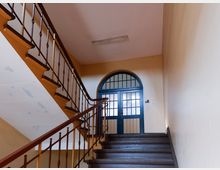 Treppenhaus in einem älteren Wohngebäude mit dunklen Holzstufen und Metallgeländer sowie Holzhandlauf. Am oberen Absatz befindet sich eine blau gestrichene Doppeltür mit halbkreisförmigem Oberlicht und strukturierten Glasfeldern.