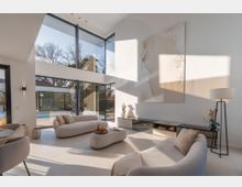 Modernes, doppelgeschossiges Wohnzimmer mit bodentiefen Glasfronten und großer Fensterfront, die auf eine Terrasse mit Pool und Garten blickt. Helle, minimalistische Einrichtung mit niedrigen Sofas, großformatigem Wandbild, flachem Sideboard und Stehlampe in neutralen Farbtönen.
