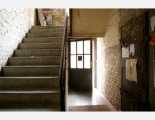 Treppenhaus mit groben Ziegelwänden, einer Metalltür mit Fensterscheiben im Hintergrund und einer Metalltür an der rechten Wand, die mit Plakaten und Zetteln beklebt ist. Oben auf der Treppe stehen Flyer und Zeitschriften in einem Wandregal.