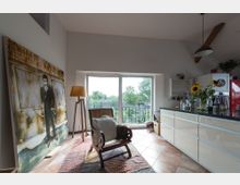 Helles Wohnzimmer mit großen Fenstern, die auf einen Balkon mit Blick ins Grüne führen. Im Raum befinden sich eine moderne Küche mit glänzenden Fronten, ein Sessel, ein großes Gemälde an der Wand und eine Stehlampe; Sonnenblumen stehen auf der Küchenarbeitsfläche.