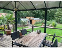 Überdachte Terrasse aus Holz mit Esstisch und Stühlen, umgeben von Topfpflanzen wie Palmen und Olivenbäumen. Im Hintergrund befindet sich ein Garten mit Rasen, einem Gewächshaus, Holzhütten und Gartenmöbeln, umgeben von dichtem Baumbestand.
