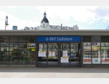 Eingang des U-Bahnhofs Südstern in Berlin mit einladendem blauen Schild über der Tür. Im Hintergrund ist ein historisches Gebäude mit Türmchen und dekorativer Fassade zu sehen. Schilder an der Eingangstür informieren über eingeschränkten Zugverkehr.
