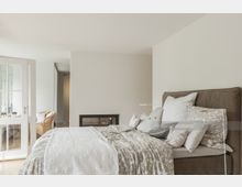 Schlafzimmer mit einem grauen Polsterbett, das mit mehreren dekorativen Kissen und einer gemusterten Tagesdecke ausgestattet ist. Im Hintergrund sind eine verglaste Doppeltür, ein Sessel aus Rattan und ein moderner Kamin erkennbar.