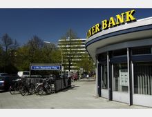 Eine Straßenszene mit dem Eingangsbereich der Berliner Bank im Vordergrund und einem U-Bahn-Schild mit der Aufschrift „U-Bhf. Bayerischer Platz“ auf der linken Seite. Im Hintergrund sind geparkte Fahrräder, Autos, Bäume und ein mehrstöckiges Gebäude zu sehen.