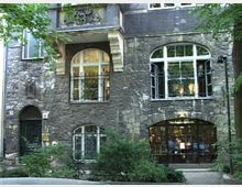 Fassade eines historischen Gebäudes aus Naturstein mit hohen, bogenförmigen Fenstern und kunstvollen Verzierungen. Im Vordergrund befinden sich Bäume und Büsche, die den Eingang und Teile der unteren Fenster umrahmen.