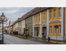 Straße mit historischen Gebäuden in einer Kleinstadt, darunter ein gelbes Gebäude mit der Aufschrift „Buchhandlung“ und „Buchdruckerei“. Die Straße ist von Blumenbeeten, Laternen und parkenden Autos gesäumt; der Himmel ist wolkenverhangen.