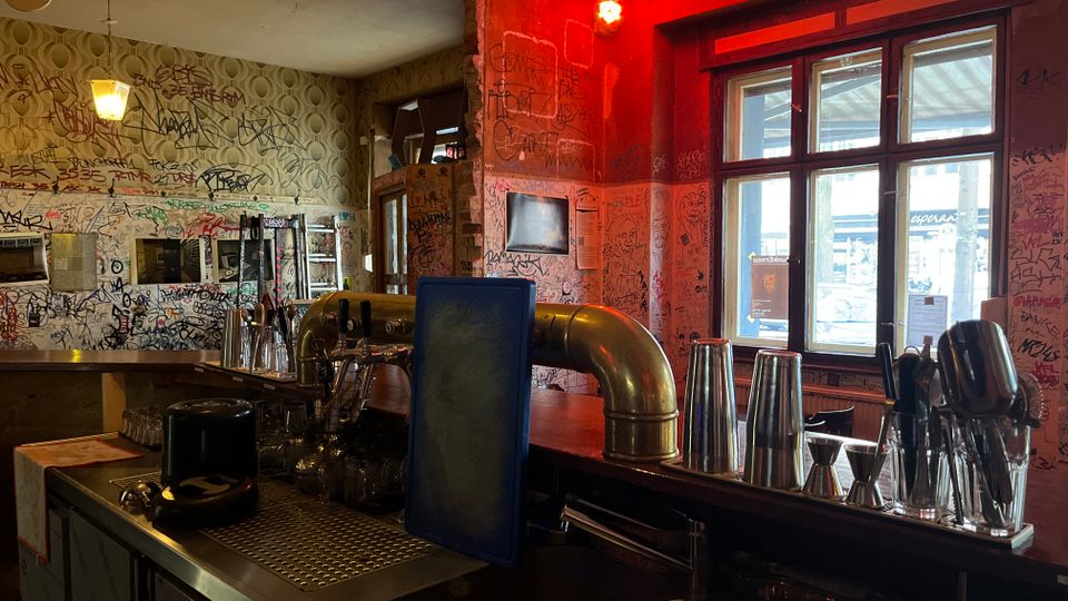 Innenansicht einer Bar mit Tresen aus Edelstahl, Gläsern und Barwerkzeugen im Vordergrund. Die Wände sind dicht mit Graffiti bedeckt, rotes Licht und eine Discokugel hängen von der Decke, daneben große Fenster zur Straße.