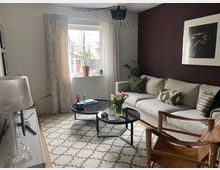 Wohnzimmer mit moderner Einrichtung: Ein helles Sofa mit dekorativen Kissen steht vor einer dunkelvioletten Wand mit einem gerahmten Kunstwerk. In der Mitte des Raumes befinden sich zwei runde, schwarze Couchtische auf einem gemusterten Teppich. Ein Fenster mit Vorhängen lässt Tageslicht herein, und Topfpflanzen schmücken den Raum.