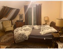 Ein gemütliches Schlafzimmer mit einem Doppelbett, geschmückt mit schwarz-weiß gemusterten Bettbezügen. Im Raum stehen ein beiger Sessel, ein Holztisch, eine Kommode, zwei Nachttischlampen und braune Vorhänge vor einem Fenster sowie einem Dachfenster; der Boden ist mit Teppichen bedeckt.