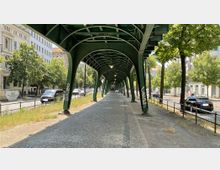 Straßenansicht mit einer Gehwegüberdachung aus grün lackierten Stahlbogenkonstruktionen unter einer Hochbahntrasse, flankiert von Bäumen, parkenden Autos und städtischer Bebauung im Hintergrund.