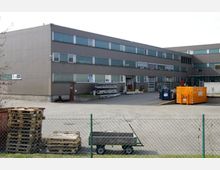 Gewerbegebäude mit grauer Fassade und mehreren Fensterreihen, umgeben von einem betonierten Hof. Im Vordergrund stehen Palettenstapel, ein Anhänger und ein Maschendrahtzaun; rechts sind große Abfallcontainer sichtbar.