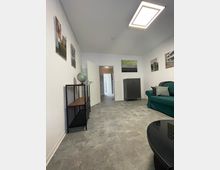 Wohnraum mit heller Wandfarbe und grauem Teppichboden. Der Raum enthält eine grüne Couch, einen schwarzen Couchtisch, einen Schrank und ein Regal mit einem Globus. An den Wänden hängen Fotografien, und im Hintergrund ist eine geöffnete Tür sichtbar, die in einen Flur führt.