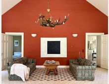 Wohnzimmer mit hoher, spitz zulaufender roter Akzentwand, großer abstrakter Wandkunst und dekorativem Kronleuchter. Zwei gemusterte Sofas stehen sich auf einem Teppich gegenüber, dazwischen ein Holztisch; seitlich führen offene weiße Doppeltüren in angrenzende Räume.