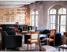 Gemütlicher Lounge-Bereich in einem rustikalen Raum mit freiliegender Holzlattenwand, großen halbkreisförmigen Fenstern und verschiedenen Sitzmöbeln, darunter Sofas, gepolsterte Stühle und Hocker. Der Boden ist teilweise mit Teppichen bedeckt und Pflanzen sorgen für Dekoration.