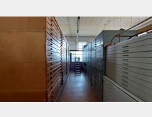 Raum mit hohen Schränken auf beiden Seiten, die aus zahlreichen flachen Schubladen bestehen, links aus Holz und rechts aus Metall. Am Ende des schmalen Gangs befindet sich ein Fenster mit einem Hocker oder Leiter davor.